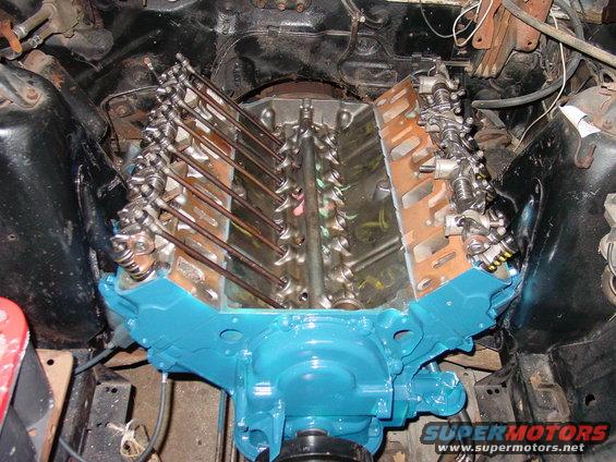 V8 engine block with valve covers removed, showing pushrods and rocker arms. The block is painted bright blue