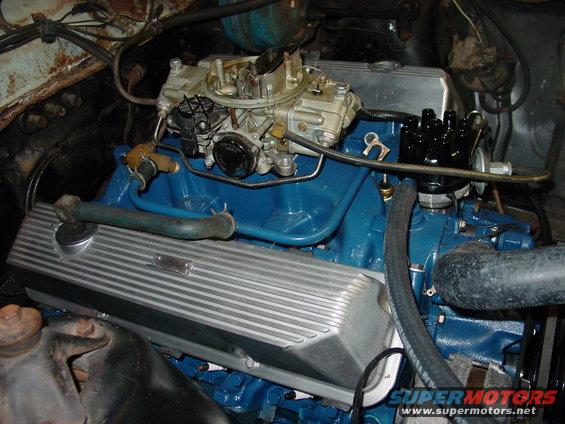 Close-up of a blue V8 engine with a carburetor and aluminum valve covers. Some rust visible on the firewall