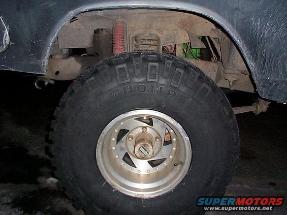 Close-up of a large off-road tire with 'THOMP' visible on the sidewall, mounted on a multi-spoke wheel. Suspension components and part of a vehicle's fender are also visible