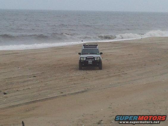 White SUV with a roof rack parked on a sandy beach near the ocean