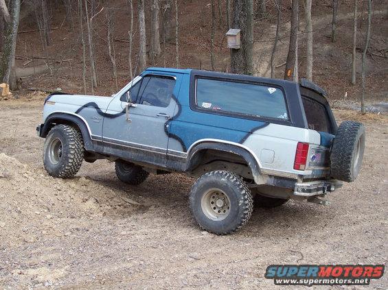 Light blue and gray Ford Bronco with aftermarket wheels and tires, parked on dirt with trees in background