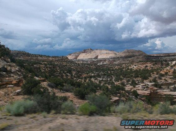 Desert landscape with rocky hills, sparse vegetation, and dramatic cloudy sky. SuperMotors logo in lower right