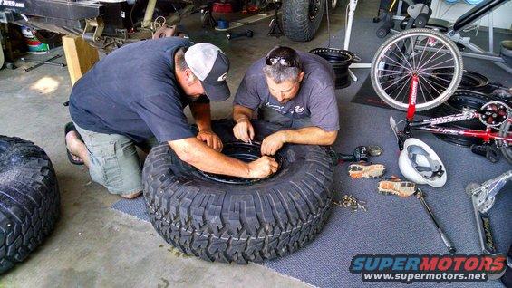 Two men kneel on a garage floor, working on a large off-road tire and rim. Tools and other tires are scattered around