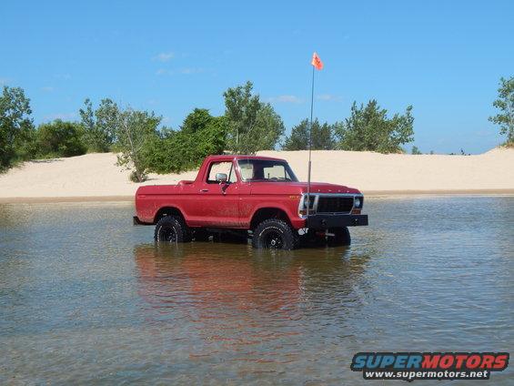 Red pickup truck driving through shallow water with sand dunes and trees in the background
