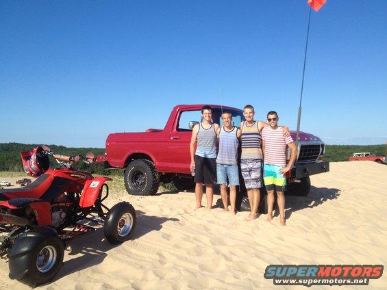 Four guys stand in front of a red Ford Bronco on a sandy dune, with an ATV nearby