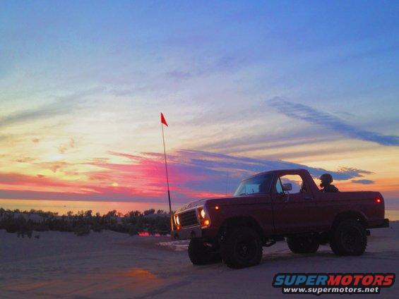 Red 1978 Ford Bronco parked on sand dunes at sunset