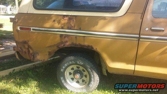 Rear side of a brown Ford Bronco with significant rust on the wheel well and lower body panel. White hardtop visible
