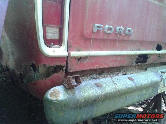 Rear of a red Ford truck with rust and damage. Shows the tailgate area and bumper