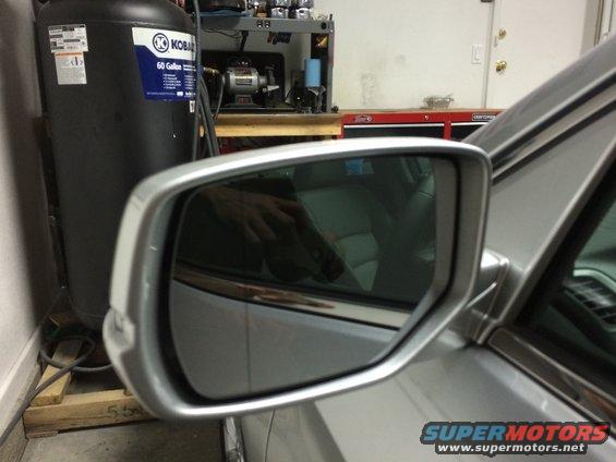 Silver car side mirror reflecting interior seats and a hand, with a large black air compressor in the background