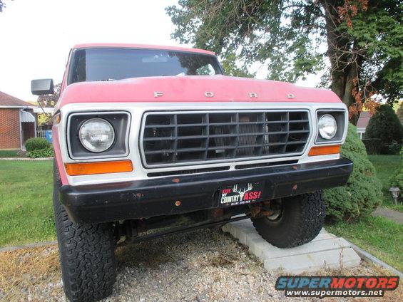 Front view of a red 1979 Ford Bronco with a black grille and a 'Kiss My Country Ass!' bumper sticker