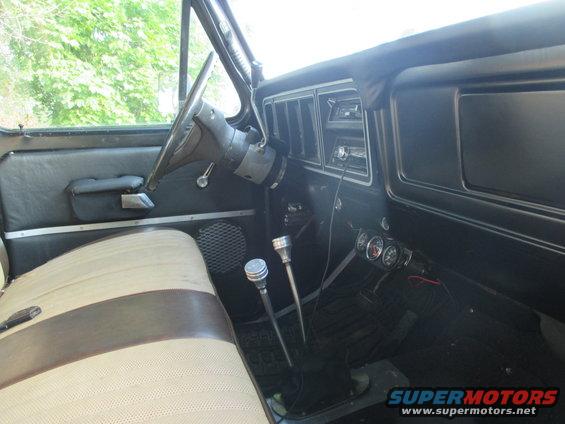1979 Ford Bronco interior showing tan and brown striped bench seat, dashboard with vents and radio, and two floor shifters