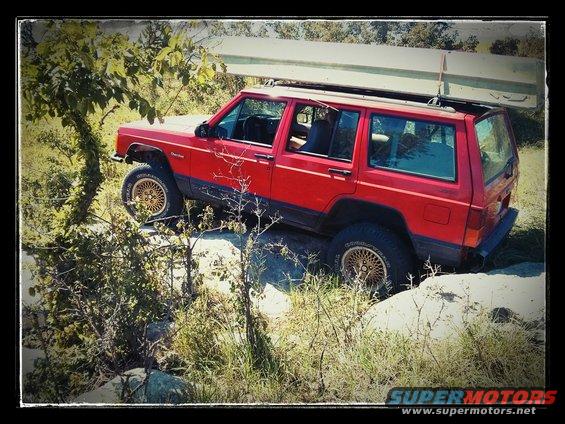 Red Jeep Cherokee XJ driving over rocks with a canoe on its roof rack
