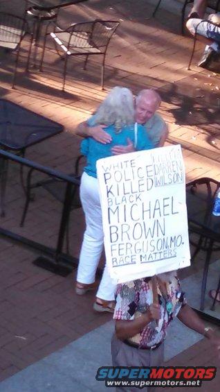 Two people embracing, one holding a sign that reads 'WHITE POLICE KILLED DARREN WILSON BLACK MICHAEL BROWN FERGUSON MO. RACE MATTERS'