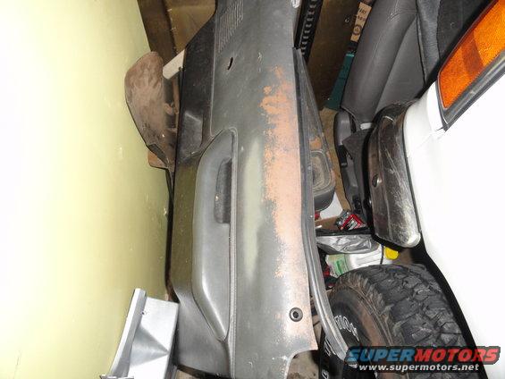 Gray car door panel with rust spots and a black handle, next to a white vehicle's fender and tire