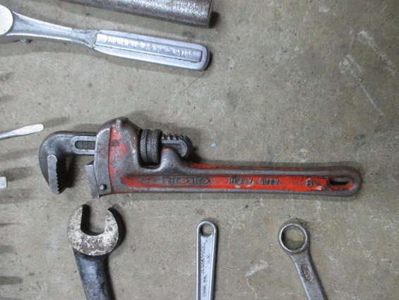 Red and silver pipe wrench with "RIDGID HEAVY DUTY" text, surrounded by other wrenches on a concrete surface
