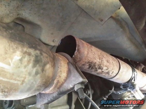 Close-up of a rusted exhaust pipe with a cut end, held by a clamp. SuperMotors.net logo visible