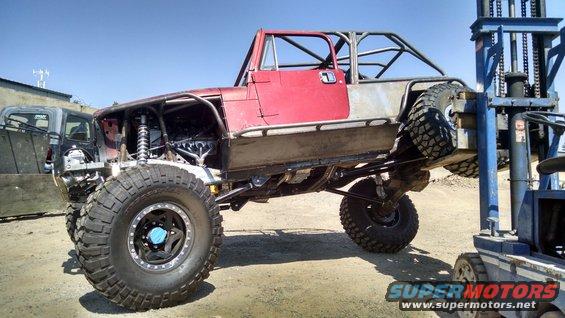 Modified Jeep CJ-style vehicle with roll cage, red doors, and large off-road tires, lifted by a forklift