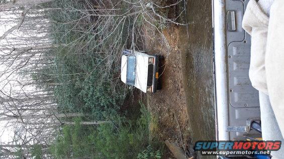 White SUV partially submerged in a creek bed, surrounded by trees