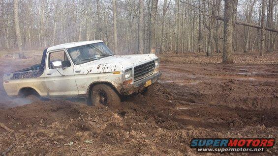 White Ford Bronco stuck in deep mud on a wooded trail. Front wheel is submerged