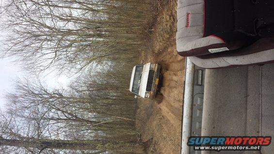White van driving on a muddy dirt road through trees. Shot from inside another vehicle