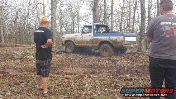 Muddy 1978 Ford Bronco with blue top stuck in wooded area, two men watching