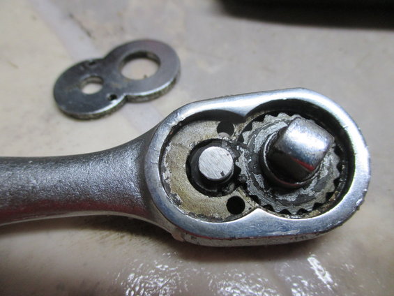Close-up of a ratchet wrench mechanism showing gears and a square drive, with a separate figure-eight shaped metal plate nearby