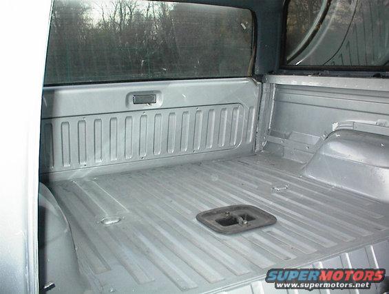 Empty silver truck bed with ribbed floor and sides. A black tie-down anchor is visible
