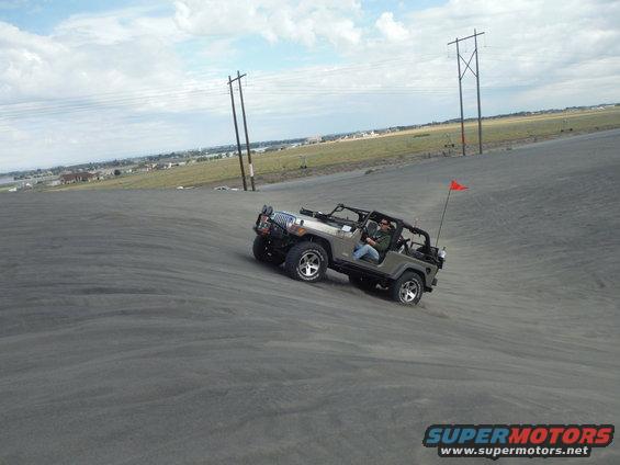 Jeep Wrangler driving up a sand dune with a person inside. Orange flag on back