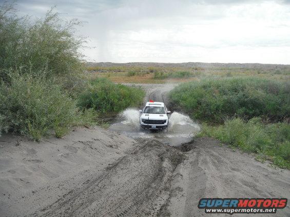 White SUV splashing through a water-filled rut on a sandy dirt track, surrounded by brush and dry grass under an overcast sky