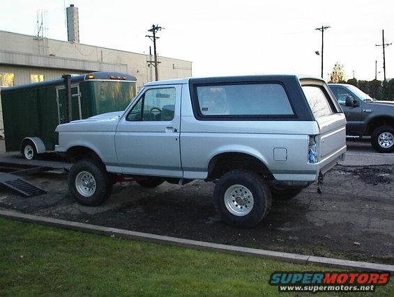 Silver Ford Bronco with black top, lifted suspension, and aftermarket wheels. Rear bumper is missing