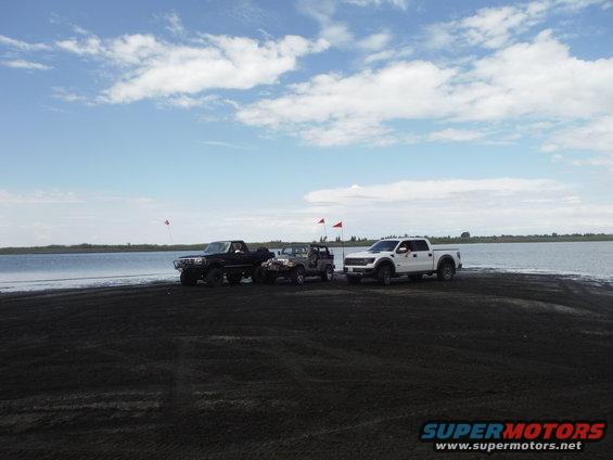 Black pickup, gray Jeep, and white pickup truck parked on dark sand near water under a cloudy sky