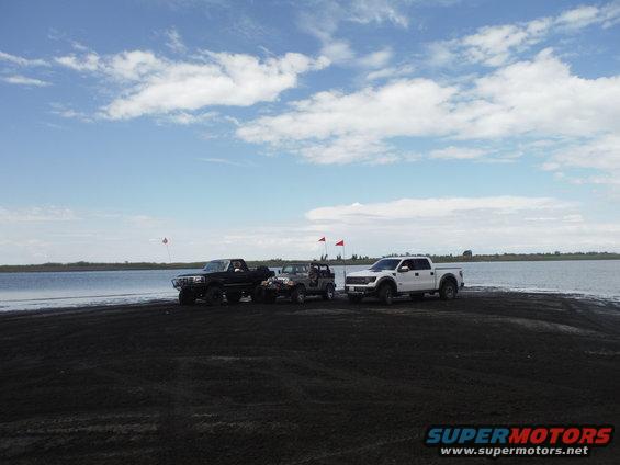 Three trucks and a Jeep parked on dark sand near a body of water under a cloudy sky