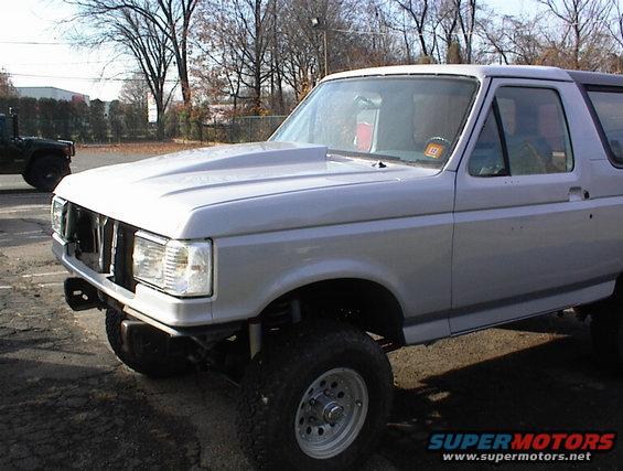 Silver Ford Bronco with a raised hood, parked outside. Another dark SUV is visible in the background