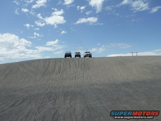 Three SUVs cresting a sand dune under a blue sky with scattered clouds
