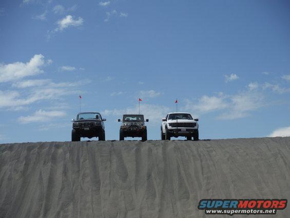 Three trucks and SUVs are lined up on top of a sand dune under a blue sky with some clouds. Each vehicle has a red flag on an antenna