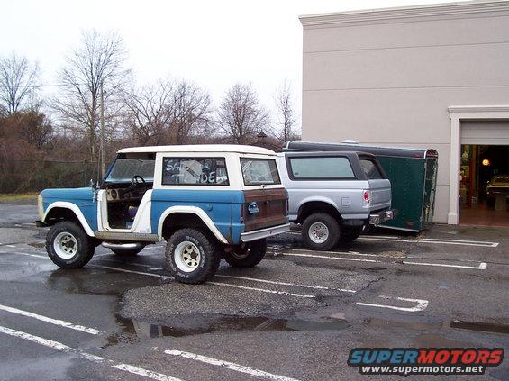 1969 Ford Bronco, blue and white, parked next to a silver Bronco and a trailer
