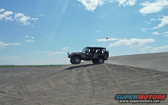 Tan Jeep Wrangler airborne over a sand dune, red flag on antenna