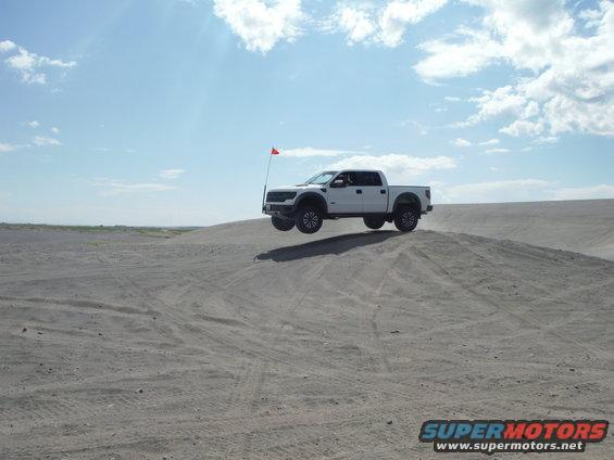 White pickup truck airborne over a sand dune with a red flag attached