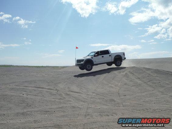 White pickup truck airborne over a sand dune, red flag visible on a pole