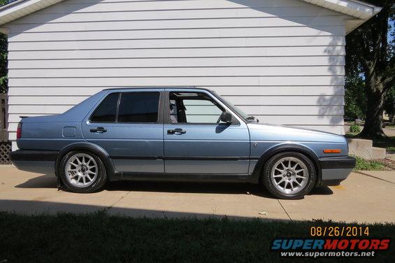 Blue 1990 Volkswagen Jetta sedan with aftermarket wheels parked in front of a white sided building