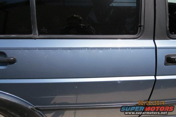Side view of a blue sedan with a pinstripe. Reflection in window shows a person