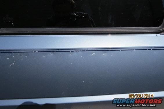 Blue car door with minor paint chips and a reflection of a person holding a camera in the window