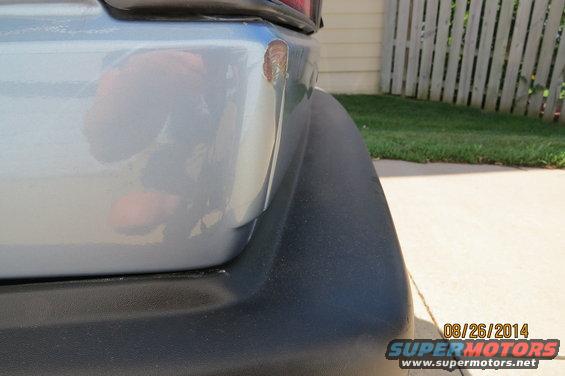Silver car's rear bumper corner with scuff and rust, black plastic trim, and a white fence in the background