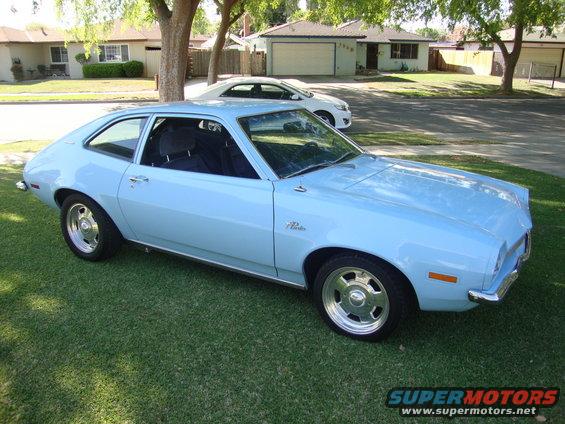 Light blue 1972 Ford Pinto coupe parked on grass in front of a suburban house. Another white car is parked in the driveway