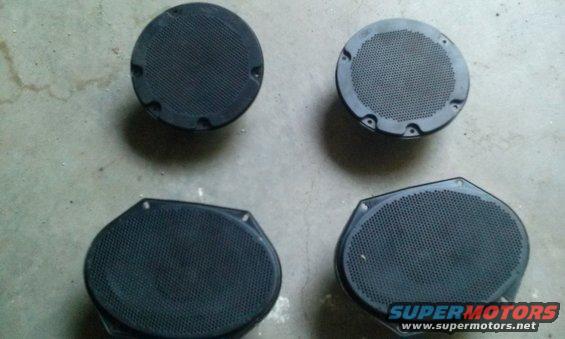 Four car speakers, two round and two oval, on a concrete surface. Supermotors.net logo visible
