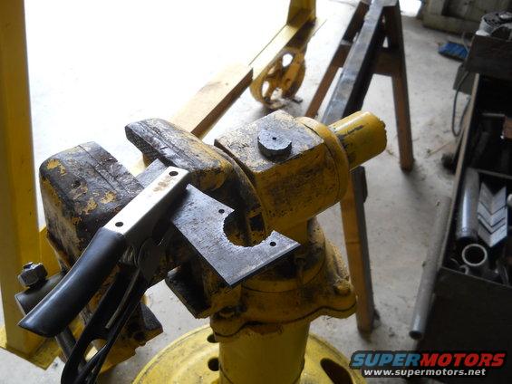 Metal plate with a cutout held in a yellow vise, being worked on with pliers