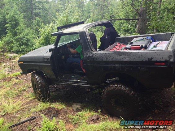 Black 1978 Ford Bronco with open doors and cargo in the bed, parked on a muddy forest trail