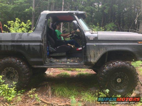 Side view of a 1979 Ford Bronco with the door open, a person inside driving on a wooded trail