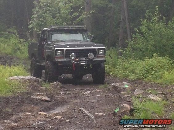 Black Ford F-Series truck with off-road tires and a light bar on top, driving on a muddy trail through trees