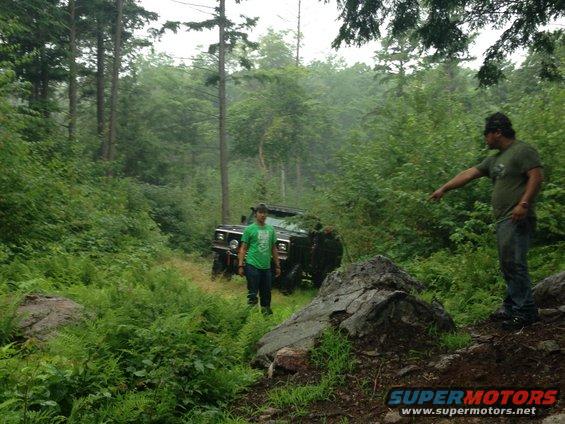 Off-road vehicle parked on a dirt path in a dense forest with two men standing nearby